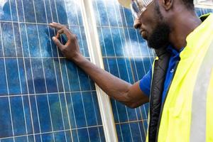 una foto al aire libre de un ingeniero africano negro inspecciona un panel solar eléctrico con casco, anteojos protectores y equipo de seguridad con una sonrisa en la cara. energía alternativa y concepto de trabajo industrial.