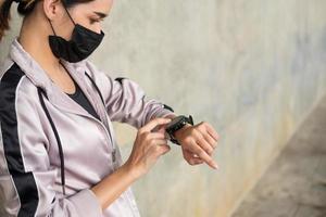 Selective focus at face of young beautiful Asian women wearing surgical face mask using smart watch to track activity before exercise or running at the park in the morning. New normal lifestyle. photo