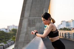 Selective focus at face of young beautiful Asian women using smart watch to track activity and listen music while warm up before exercise or running at the urban city view. Fit and healthy lifestyle. photo