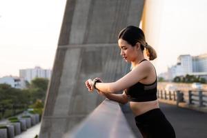 Selective focus at face of young beautiful Asian women using smart watch to track activity and listen music while warm up before exercise or running at the urban city view. Fit and healthy lifestyle. photo