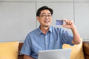 tiro interior dentro de la casa de los hombres asiáticos mayores sentarse en el sofá y usar la computadora portátil para el pago de la tarjeta de crédito en línea con una sonrisa en su rostro. senior feliz con la tecnología. foto