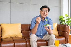 hombres asiáticos mayores sentados en el sofá de casa mientras comen ensalada de vegetales verdes y jugo de naranja orgánico para el desayuno. estilo de vida de alimentación saludable para el concepto de personas mayores. foto