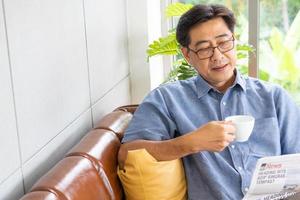 Selective focus at face. Senior Asian man reading newspaper mock up at the sofa couch while drinking coffee in the morning. Happy retirement with relaxation lifestyle and routine. photo