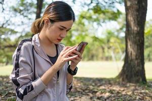 enfoque selectivo en la cara de las jóvenes y hermosas mujeres asiáticas que usan un reloj inteligente para rastrear la actividad y escuchar música desde el teléfono inteligente mientras se calientan antes de hacer ejercicio de yoga con árboles naturales y de fondo. foto