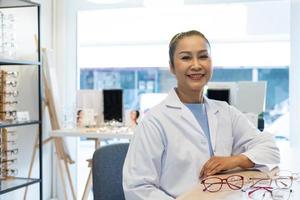 Asian women Doctor optometrist smiling while using tablet and recommend customer to choose glasses beautiful frame trial inside of optical shop, store. Eye sight for patient, healthcare concept. photo