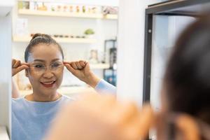 enfoque selectivo en el espejo. Las ancianas asiáticas mayores sonríen y miran el espejo mientras eligen hermosas gafas de prueba dentro de la tienda óptica, tienda. vista para ancianos, atención médica. foto