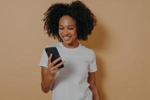 Happy african female student using mobile phone while standing isolated over sand color background photo