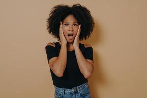 Photo of shocked pretty dark skinned curly haired lady holding hands on cheekbones