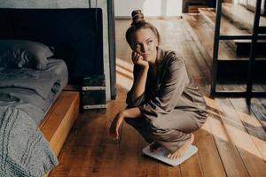 Upset young attractive woman using scales at home and wondering why cannot lose weight photo