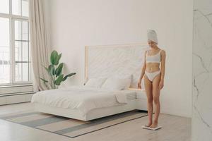 Shot of healthy young slender woman in underwear standing on scales in bedroom photo