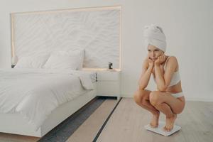 Young sad beautiful woman in white underwear standing on electronic smart scales in bedroom photo