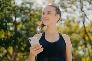 Thoughtful cheerful woman with pony tail has walk outdoor listens music while going in for sport holds modern smartphone leads healthy lifestyle focused into distance poses at park against trees photo