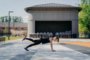 Sportive young woman being in good physical shape poses in plank raises leg wears sportsclothes listens music via wireless earphones breathes fresh air goes in for sport regularly on stadium photo