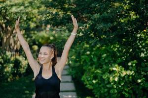 Photo of cheerful young woman with smile on face keeps arms raised up being in good mood dressed in active wear poses against green nature background. People healthy lifestyle recreation concept