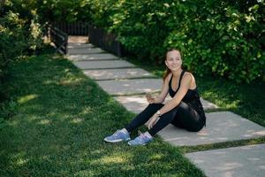 Outdoor shot of active brunette European woman downloads music on smartphone for active training outside has break after workout dressed in active wear shares content in blog looks happily away photo