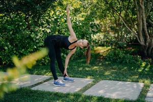 Active fitness model exercises outdoors leans to feet raises arm prepares muscles before cardio training challenges herself dressed in active wear has pony tail poses in green park during sunny day photo