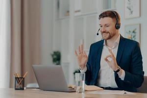 Career and distance job concept. Pleased bearded man corporate worker dressed formally gestures okay sign at webcam of laptop uses headset for online calling poses at desktop gains new knowledge photo