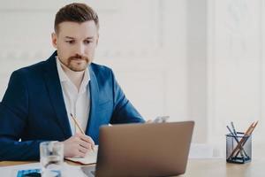 Serious bearded man marketer involved in working process makes notes with pencil looks attentively at laptop computer wears blue formal suit poses at coworking space writes organisation plan photo