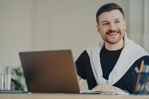 Happy bearded man office worker writes organisation plan works remotely makes notes of info sits in front of opened laptop looks away dressed in casual t shirt and jumper tied over shoulders photo