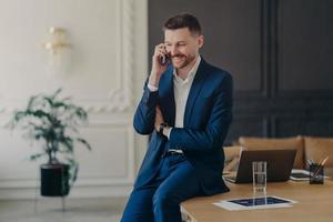 Smiling executive manager talking on phone while sitting on table in office photo