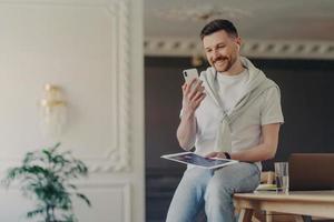 Indoor shot of bearded adult man freelancer poses in coworking space makes video call via smartphone uses wireless earphones connected to free high speed internet has online conference with coworkers photo