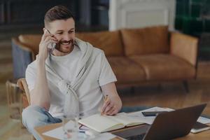 Cheerful man freelancer makes telephone call writes down information in notepad makes notes in planner looks attentively at laptop screen poses against home interior at desktop. Technology user photo