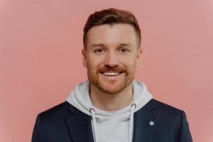 Portrait of cheerful European man with stubble dressed in hoodie and black jacket looks directly at camera has happy face expression poses against pink background. People emotions lifestyle. photo