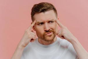 Close up shot of serious bearded adult man keeps fingers on temples concentrated aside tries to recall something in mind dressed casually isolated over pink background. Male model thinks deeply photo