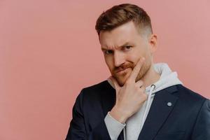 Portrait of serious man holds chin looks displeased at camera being deep in thoughts wears stylish clothes tries to make decision isolated over pink background with blank space for your promo photo