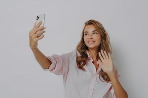 Glad young European woman waves palm shows hello gesture poses at smartphone camera photo