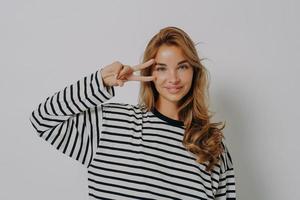 jovencita alegre y optimista con blusa a rayas que muestra el signo de la paz con los dedos, aislada en una pared gris foto