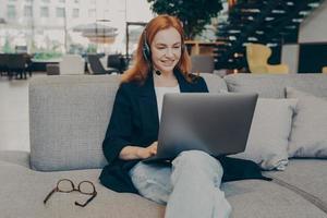 Smiling lady freelancer wearing wireless headset, communicating with client via video call on laptop photo