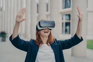 mujer de negocios de jengibre impresionada probando la realidad virtual, usando auriculares vr móviles afuera foto
