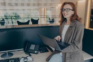 Woman in casual clothes sitting on desk in her cubicle in office while holding laptop and smiling photo
