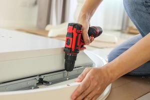 man assembling white table furniture at home using cordless screwdriver photo