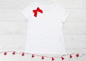 Close up white blank template t shirt with copy space and Christmas Holiday concept. Top view mockup t-shirt and red bow on white wooden background. Happy New Year decorations accessories. Xmas outfit photo