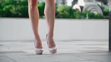 mulher de negócios de sucesso sexy em um vestido rosa andando no parque da cidade. belas pernas femininas de salto alto estão andando por uma rua urbana contra o pano de fundo da luz do sol. câmera lenta. video