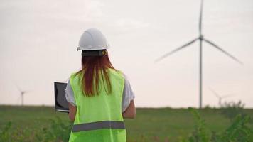 mujer especialista en ecología en el control uniforme con el funcionamiento correcto de la computadora portátil y la eficiencia de los molinos de viento al atardecer. Ingeniera monitoreando el rendimiento del sistema y tomando notas en el programa. video