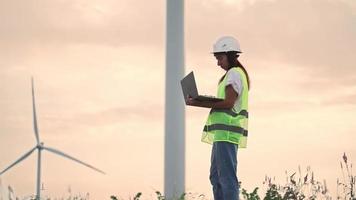 Woman Ecology Specialist in Uniform Checking with Laptop Correct Operation and Efficiency Windmills on Sunset. Female Engineer Monitoring System Performance and Making Notes in Program. video
