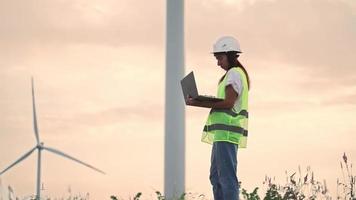 donna ecologia specialista nel uniforme controllo con il computer portatile corretta operazione e efficienza mulini a vento su tramonto. femmina ingegnere monitoraggio sistema prestazione e fabbricazione Appunti nel programma. video