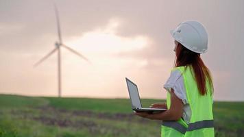 donna ecologia specialista nel uniforme controllo con il computer portatile corretta operazione e efficienza mulini a vento su tramonto. femmina ingegnere monitoraggio sistema prestazione e fabbricazione Appunti nel programma. video