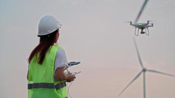engenheira de ecologia de mulher de uniforme e capacete segurando joystick controlando drone voador trabalhando no moinho de vento no belo fundo por do sol. alternativa à energia elétrica. video