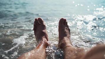 Close-up on the legs of an unrecognizable man relaxing on the sea beach on a summer sunny day. Vacation and travel concept. From the first person. POV video