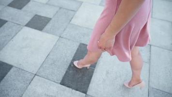 sexy erfolgreiche Geschäftsfrau in einem rosafarbenen Kleid, das im Stadtpark spazieren geht. schöne weibliche beine in high heels gehen vor dem hintergrund des sonnenuntergangslichts eine städtische straße entlang. Zeitlupe. video