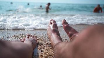 una pareja casada irreconocible descansa en la playa cerca del mar. primer plano de piernas masculinas y femeninas chapoteando en agua de mar. camara lenta. video