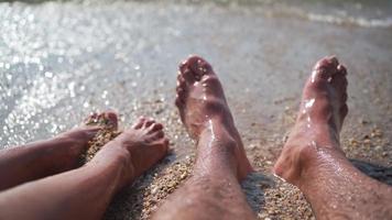 irriconoscibile sposato coppia è riposo su il spiaggia vicino il mare. avvicinamento di maschio e femmina gambe spruzzi nel mare acqua. lento movimento. video