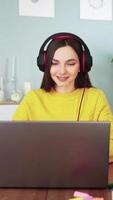 mujer feliz en auriculares comprometidos e learning web cam chat home. una joven estudiante con suéter amarillo en una laptop se comunica con videollamadas en línea, educación a distancia y tecnología moderna. vídeos verticales video