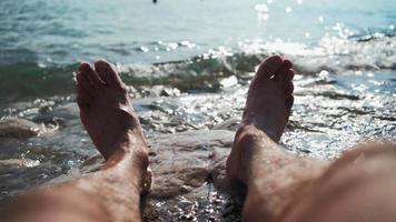 close-up nas pernas de um homem irreconhecível relaxando na praia do mar em um dia ensolarado de verão. conceito de férias e viagens. da primeira pessoa. pov video