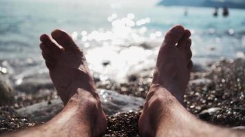 Close-up on the legs of an unrecognizable man relaxing on the sea beach on a summer sunny day. Vacation and travel concept. From the first person. POV video