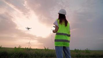 femme ingénieur en écologie en uniforme et casque tenant le joystick contrôlant le drone volant travaillant au moulin à vent sur fond de coucher de soleil magnifique. alternative à l'énergie électrique. video
